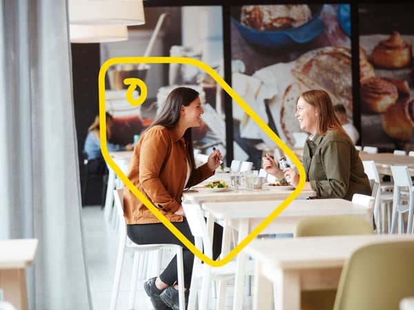 Two smiling women sitting at a white table eating a meal together in the IKEA restaurant, with a food poster in the background.
