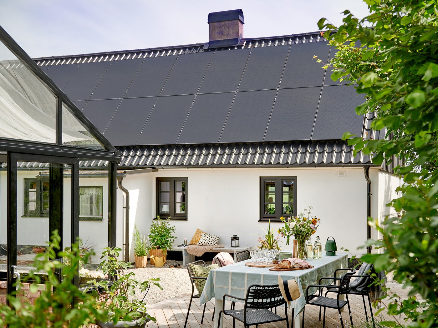 Ett stort vitt hus med solpaneler på taket. Framför huset finns en uteplats med utemöbler och grönska. 