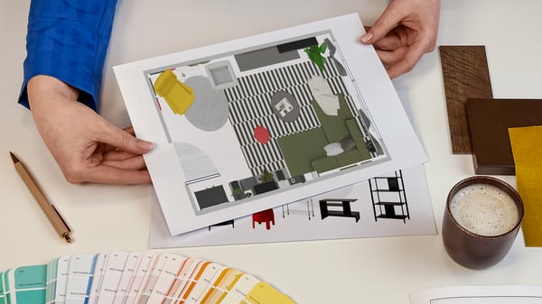Projektantka wnętrz IKEA prezentująca kartki z wydrukiem rzutu z góry zaprojektowanego pomieszczenia. Obok na blacie leży wzornik kolorów i próbki materiałów.