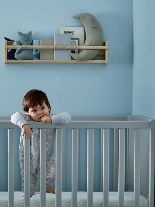 A grey cot with a child inside, leaning on one of the sides. A beige wall storage with items is behind the child.
