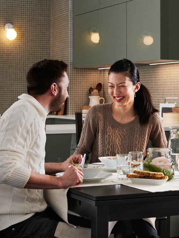 A man and a woman are sitting at a table in a kitchen enjoying a meal. They are both laughing.