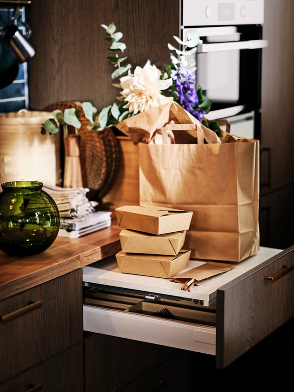 A green KONSTFULL vase on a worktop next to an open UTRUSTA pull-out work surface with a brown paper bag on top.