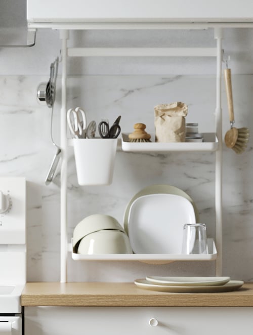 Close-up of a white kitchen organizer set with pairs of scissors, bowls, glasses, and plates, placed above a beige worktop. 