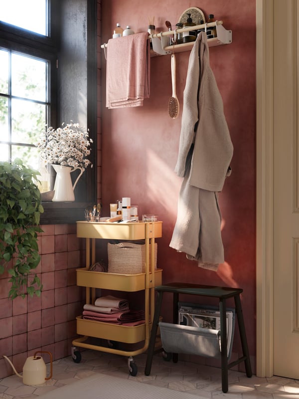 Un baño rosa tiene un carrito RÅSKOG amarillo con artículos de baño, una toalla y una bata encima sobre rieles y ganchos.