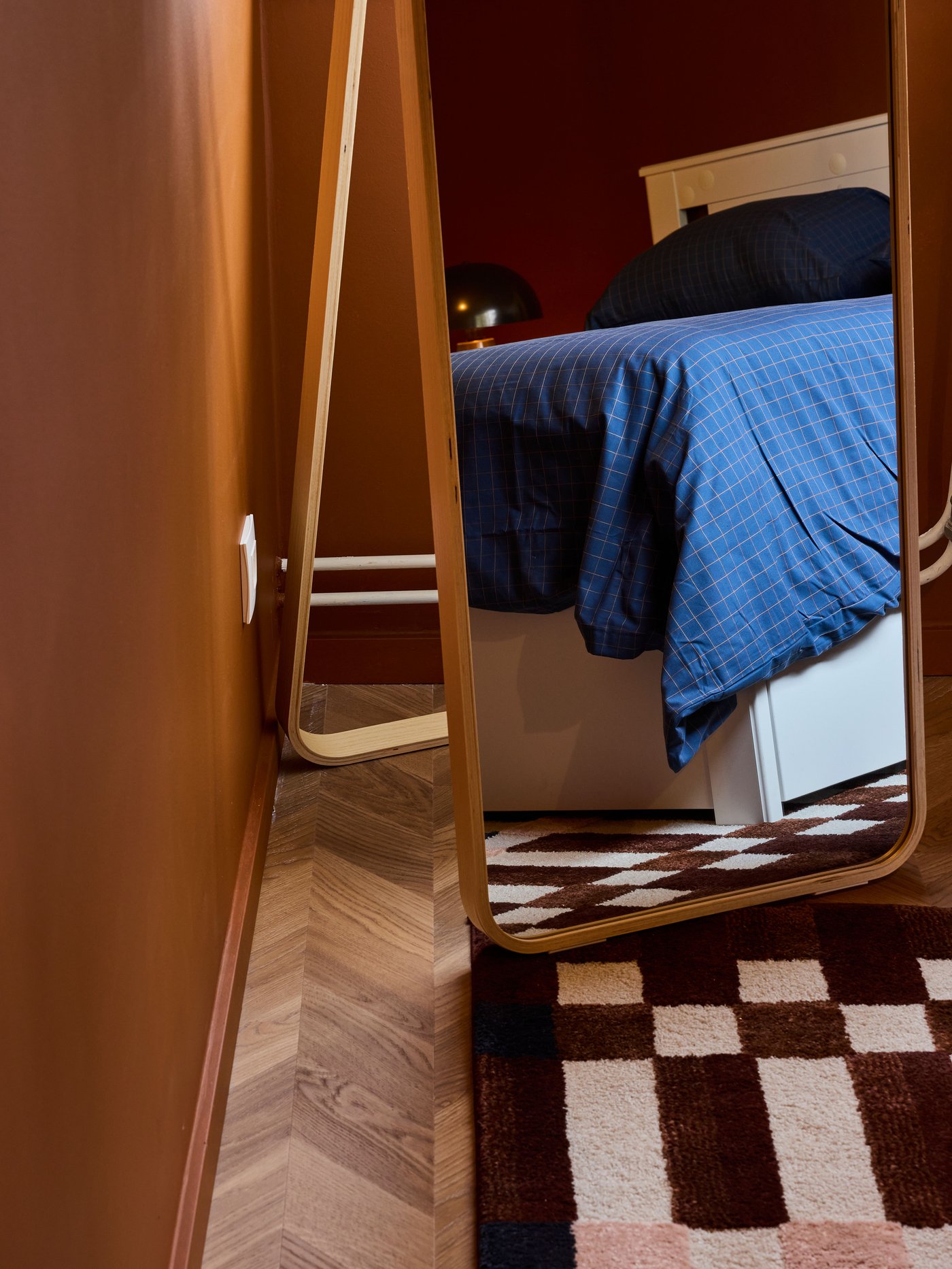Staande spiegel naast bed met blauw geruit dekbedovertrek, op een bruin‑wit tapijt in een warme slaapkamer met houten vloer.