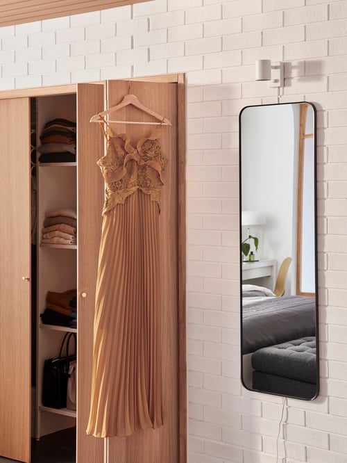A LINDBYN mirror with rounded corners is placed next to an AURDAL wardrobe. On the door is a dress on a BUMERANG hanger.
