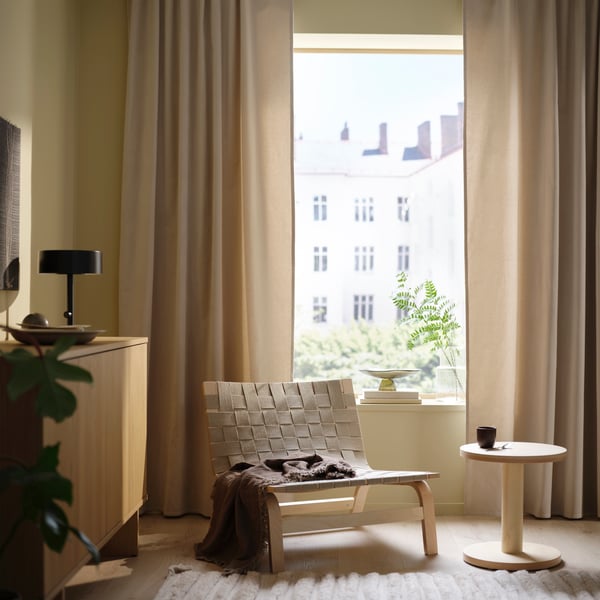 A bright, minimalist living space with beige curtains, a woven lounge chair, a small round side table with a cup, and a wooden cabinet with a black lamp. Sunlight streams through the large window, highlighting a potted plant on the windowsill.