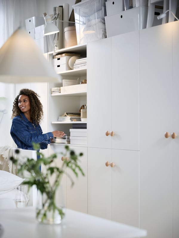 Een vrouw met krullend haar, in spijkerjack, organiseert haar lichte IKEA LASTARE opbergoplossing: witte modulaire kasten, witte KUGGIS en transparante SAMLA dozen, met houten knoppen.