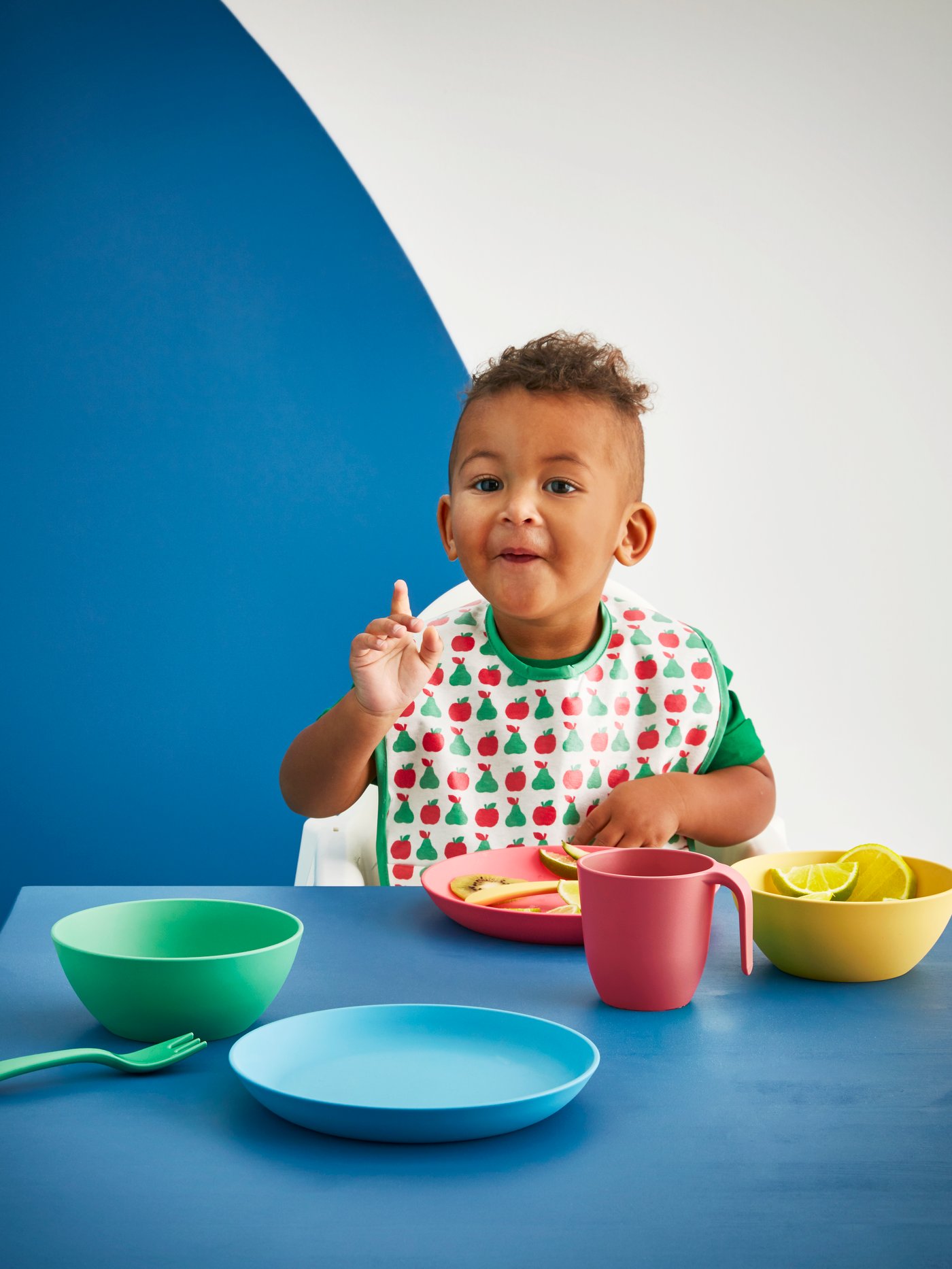 Seorang budak lelaki memakai alas dada MATVRÅ dalam kerusi tinggi di meja dengan mangkuk HEROISK, pinggan, koleh dan kutleri.