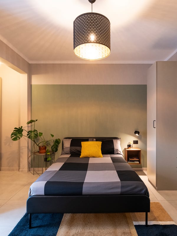 Un camera con letto al centro, tavolino vassoio GLADOM con pianta sopra e un mobile ENHET a parete ai lati. Sulla parete di destra sono appoggiati un mobile alto e un attaccappani TJUSIG su cui sono appoggiate due giacche e una borsa. Sotto il letto un tappeto a fantasie geometriche.
