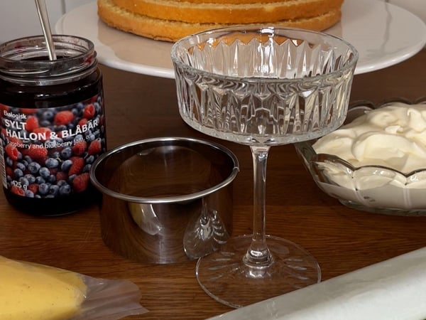 En närbild på en ekologisk SYLT HALLON & BLÅBÄR hallon- och blåbärssylt, en LÄTTBAKAD kakskärare och ett SÄLLSKAPLIG glas.