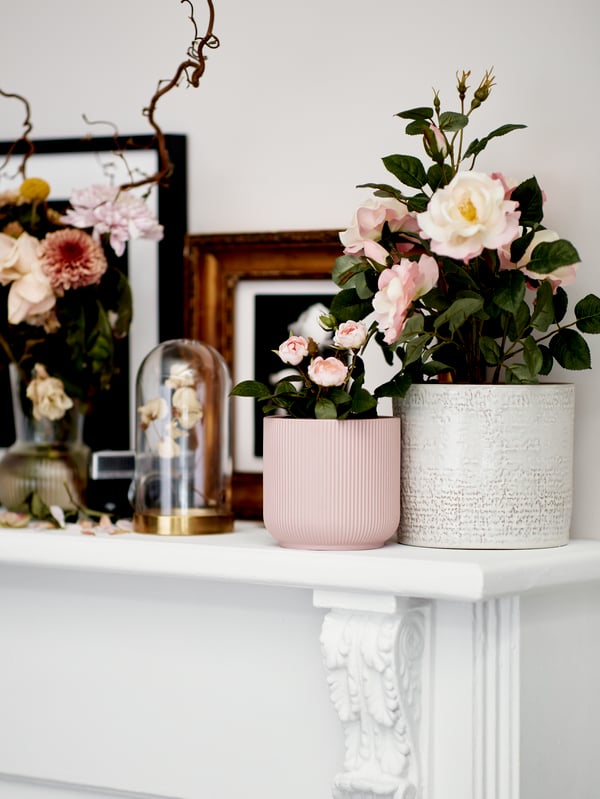 A white table placed against a wall is filled with decorations, including flowers in a small pink GRADVIS plant pot and a bigger CHIAFRÖN plant pot in white; dried flowers placed in a BEGÅVNING glass done with a gold base; and various picture frames.