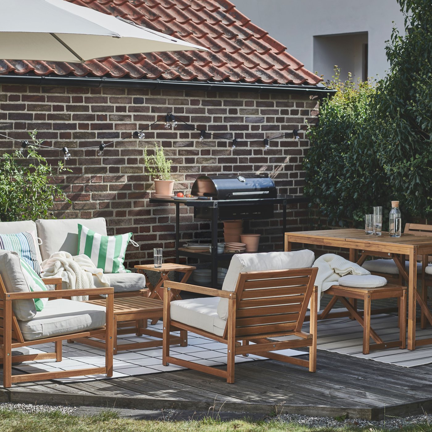 Es ist ein sonniges Holzdeck vor einer Backsteinwand zu sehen. Darauf sind ein NÄMMARÖ Tisch, Sitzgelegenheiten und ein GRILLSKÄR Grill platziert.