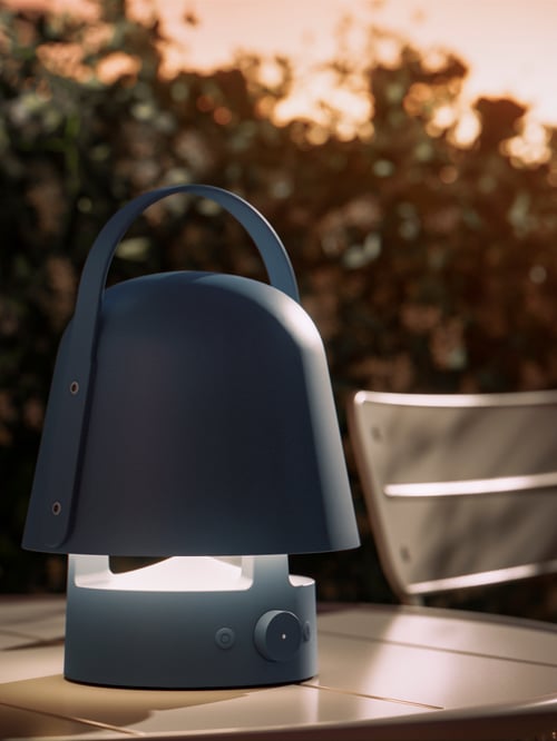 A close-up of the blue VAPPEBY Bluetooth speaker lamp perched on an outdoor table at evening; the lamp is on.