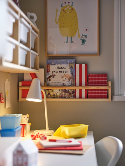 Children's room with a white desk with pull-out storage, a lit work lamp, a yellow box, a framed drawing, and wall storage.