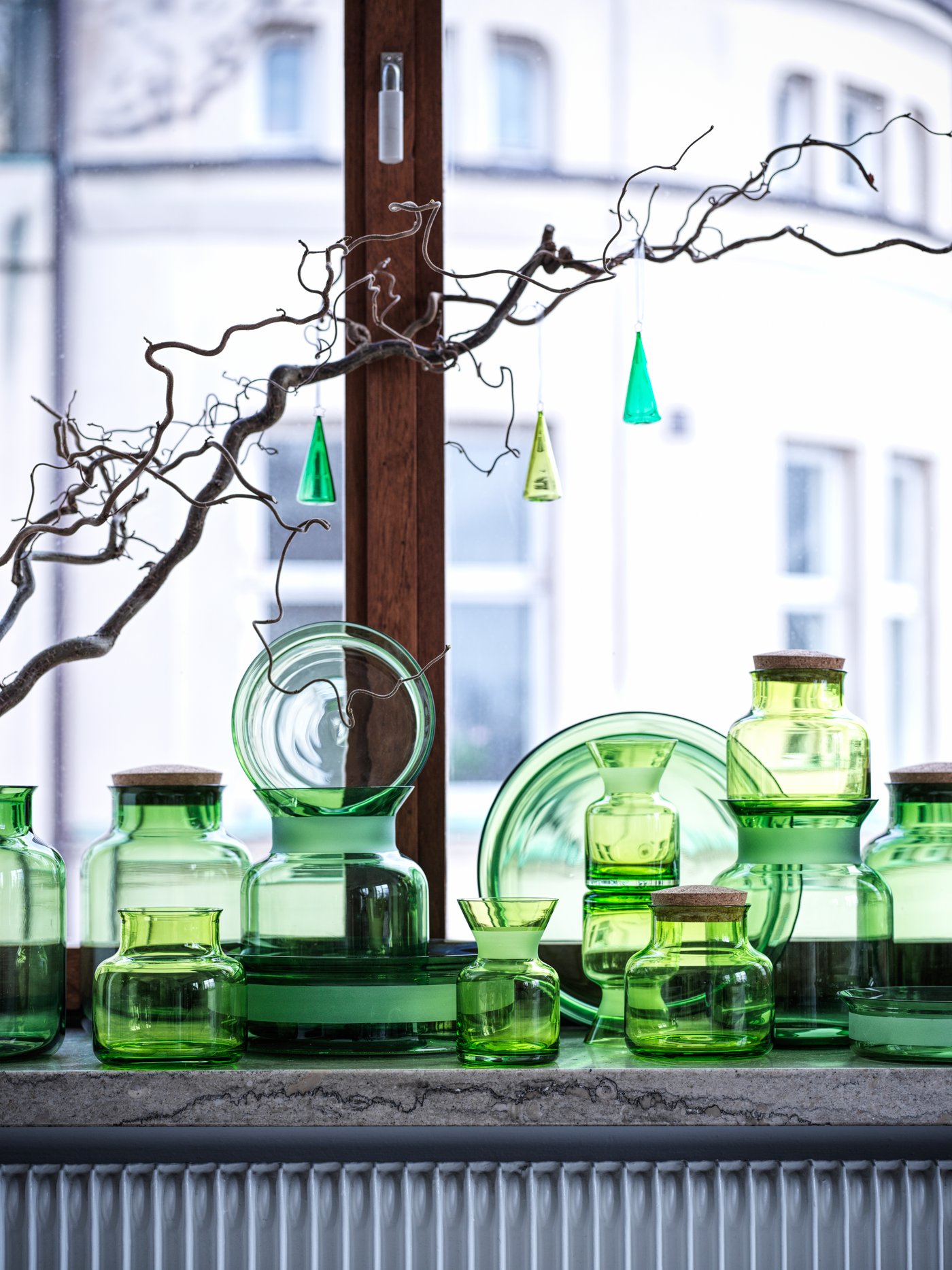 Set of three green VINTER 2021 hanging spruce tree-shaped decorations hang from a branch in a window above glass vases.