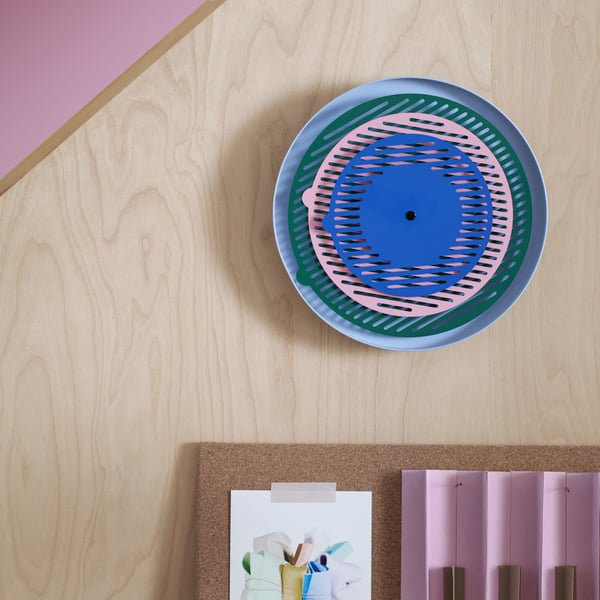 A colourful TESAMMANS wall clock, with green, pink and blue circles that move to show the time, hangs on a wooden wall.