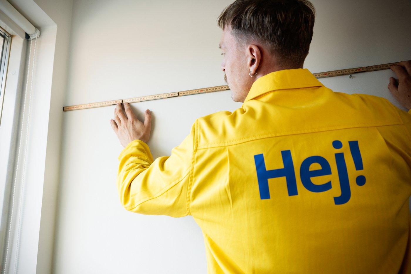 Person i gul arbetsjacka mäter en vägg med tumstock nära ett fönster.