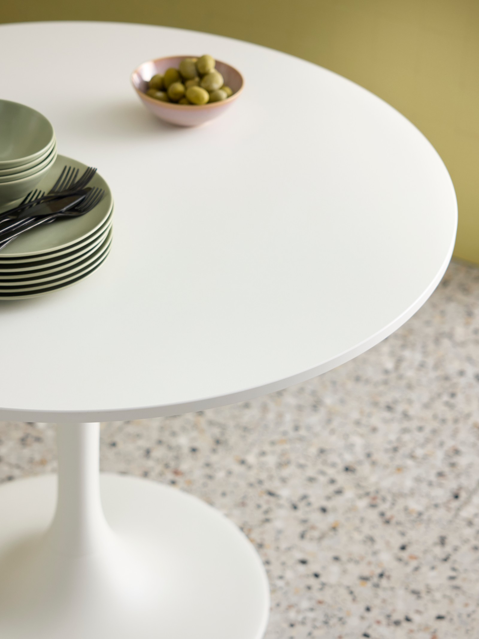 Close up of the white, round DOCKSTA table with green plates and a small bowl of olives on it. Placed on a beige carpet.