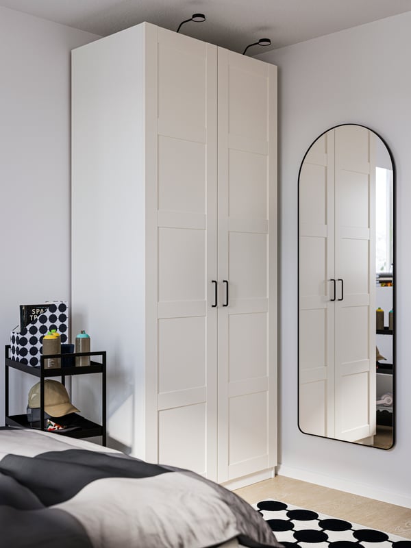 Armoire-penderie PAX blanche dans le coin d’une pièce, avec un miroir LINDBYN sur le mur à droite et un lit au premier plan.