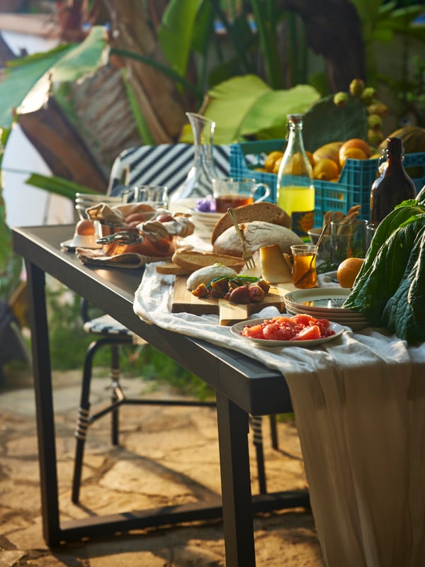 Una mesa VÄRMANSÖ preparada para una fiesta de verano con frutas, verduras y bebidas en una botella KORKEN.
