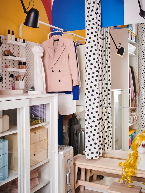 A storage area with a white SYVDE cabinet with boxes inside, clothes rails with hanging clothes and a GO mirror on the wall.