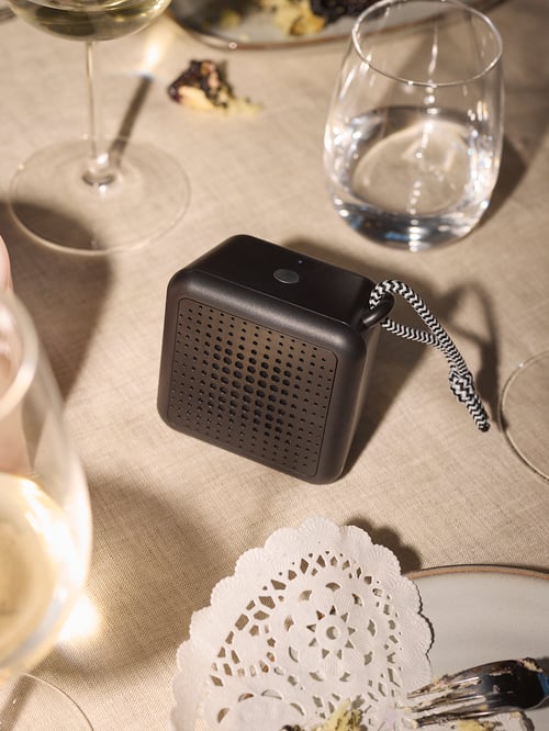 Overhead shot of a black VAPPEBY Bluetooth speaker on a table surface, set between a GLADELIG side plate and a glass.