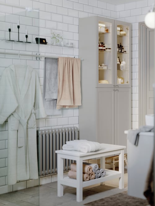 The corner of a tiled bathroom with towels and a bathrobe on a towel rail on the wall beside two GODMORGON high cabinets.