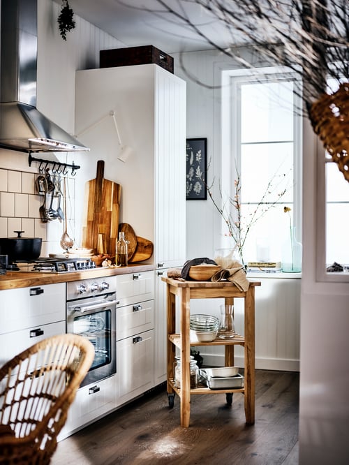 A white country-style kitchen with a wooden worktop and white cabinets, and a BEKVÄM trolley in the middle of the room.