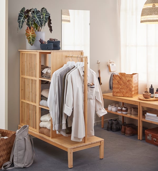 A NORDKISA open wardrobe made of bamboo with folded and hung clothes is placed in front of a brown wall, beside a mirror and shoe racks.