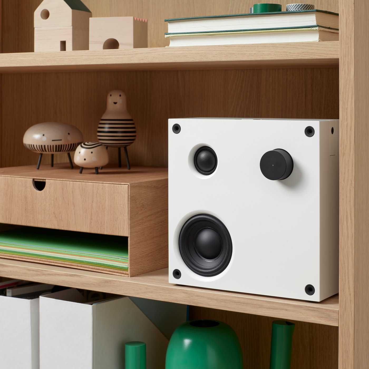 A square, white VAPPEBY Bluetooth speaker with black details like knobs and screws, is on a wooden shelf by decorative items.