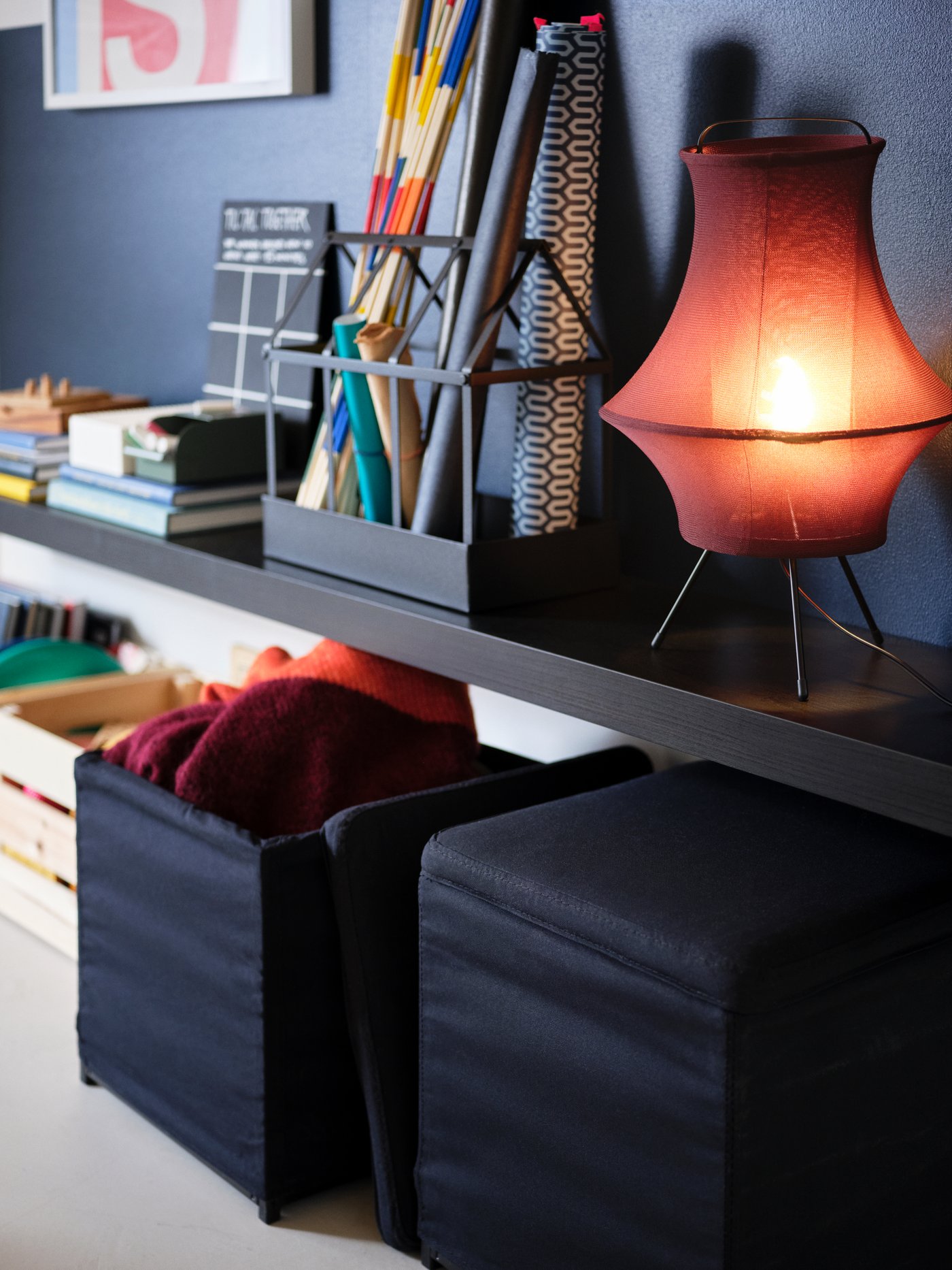 Dark red FYXNÄS table lamp and various items on a black-brown LACK wall shelf above two black BOSNÄS footstools with storage.