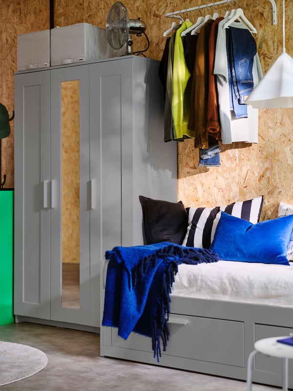 A grey BRIMNES wardrobe with 3 doors beside a BRIMNES day-bed with a blue KLIPPOXEL throw and various cushions on top.