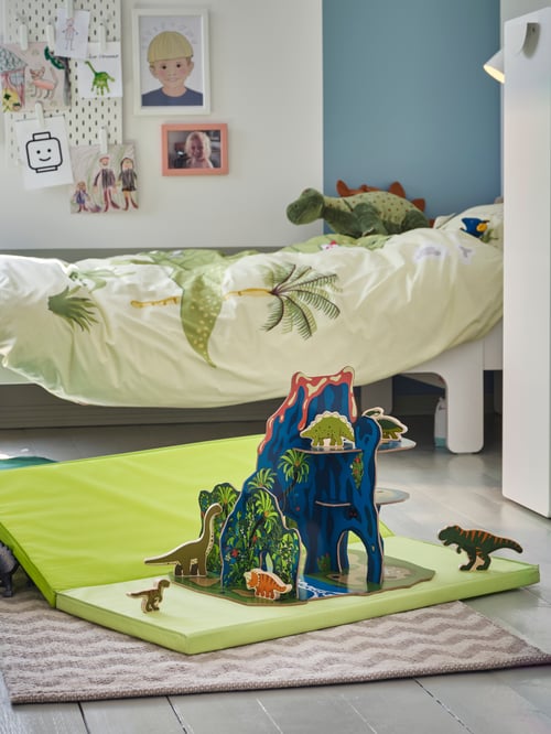 Children's room with a folding gym mat on which are dinosaur toys, a rug, a green duvet set, and a wall-mounted pink frame.