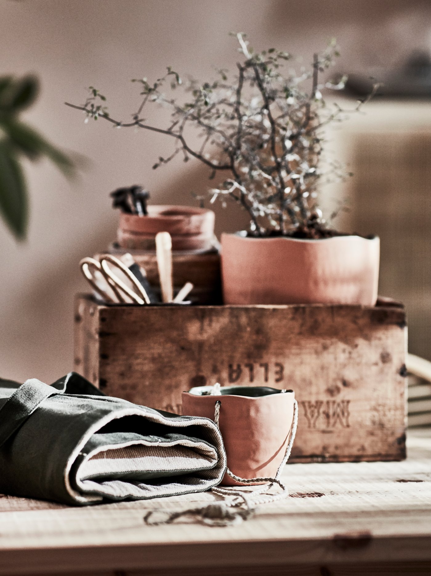 A terracotta hanging planter on a wooden table. There is a potted plant and gardening accessories in a box behind it.