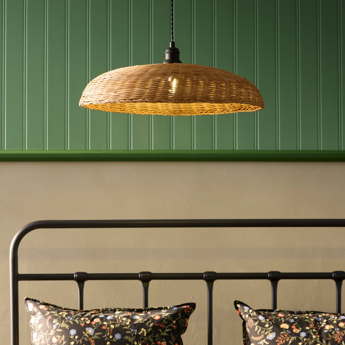 A rattan NARRKOLV pendant lamp hangs from the ceiling above a stylish bed with floral pillows set against a green wall.