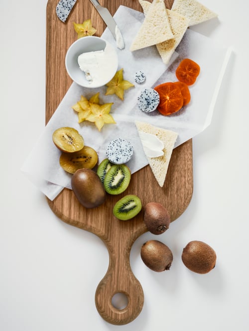 An ARTISTISK chopping board with fruits like kiwis and star fruit placed together with butter and triangle slices of bread.