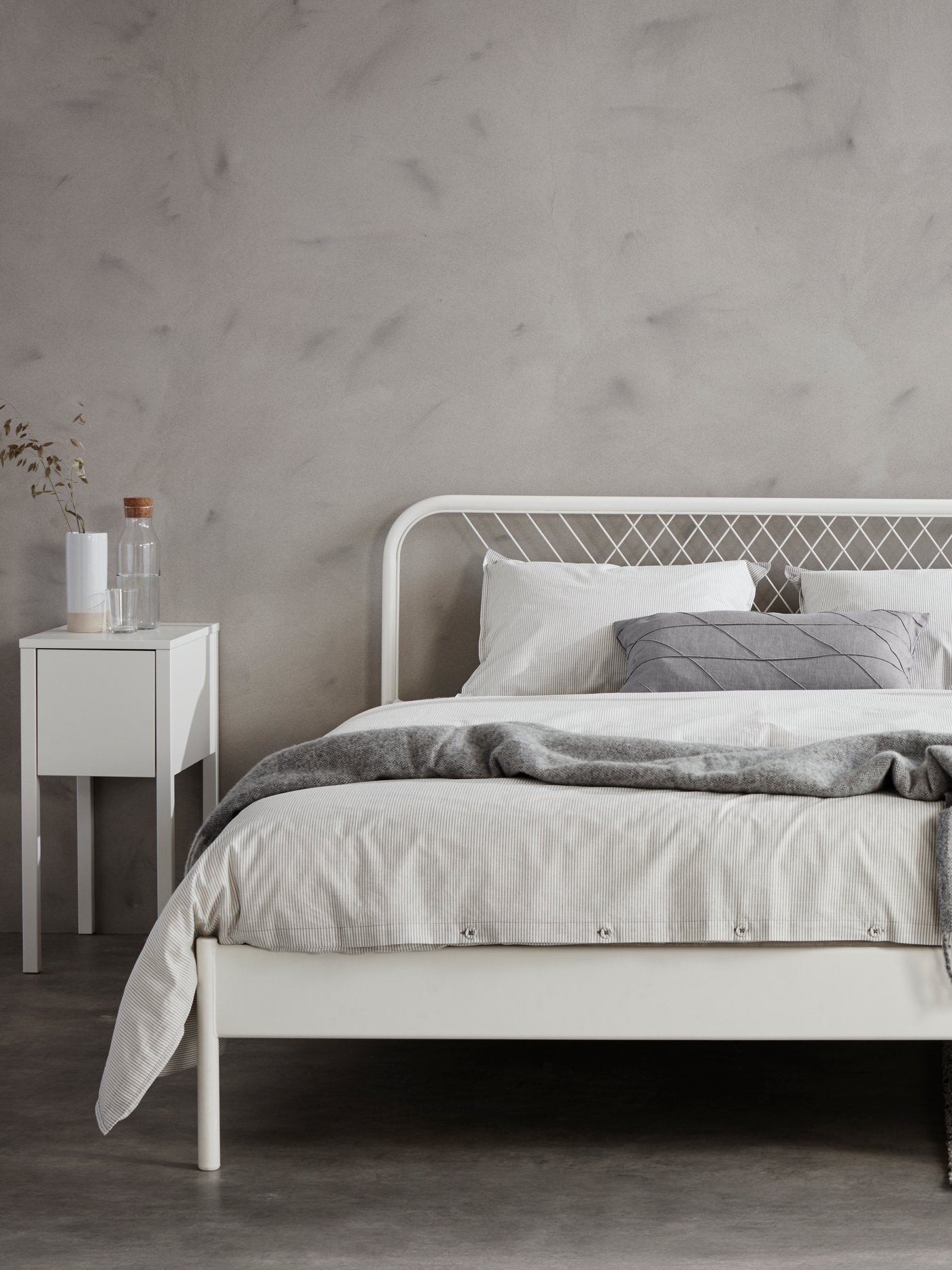 Bedroom setting with a bed frame with mattress and duvet cover with three cushions, a bedside table, and carafe with stopper.