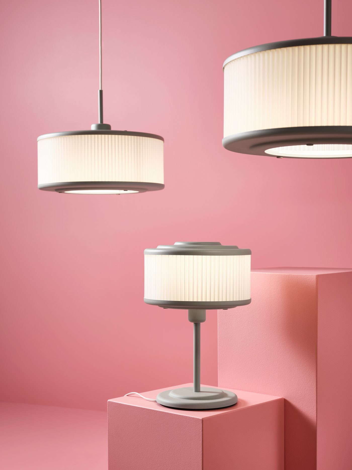 Two REMTÅNG pendant lamps hang in a light pink space. A REMTÅNG desk lamp in placed on a pink pedestal.