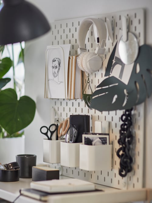 Close-up of white SKÅDIS pegboard with stationery containers, headphones and drawings attached to it.