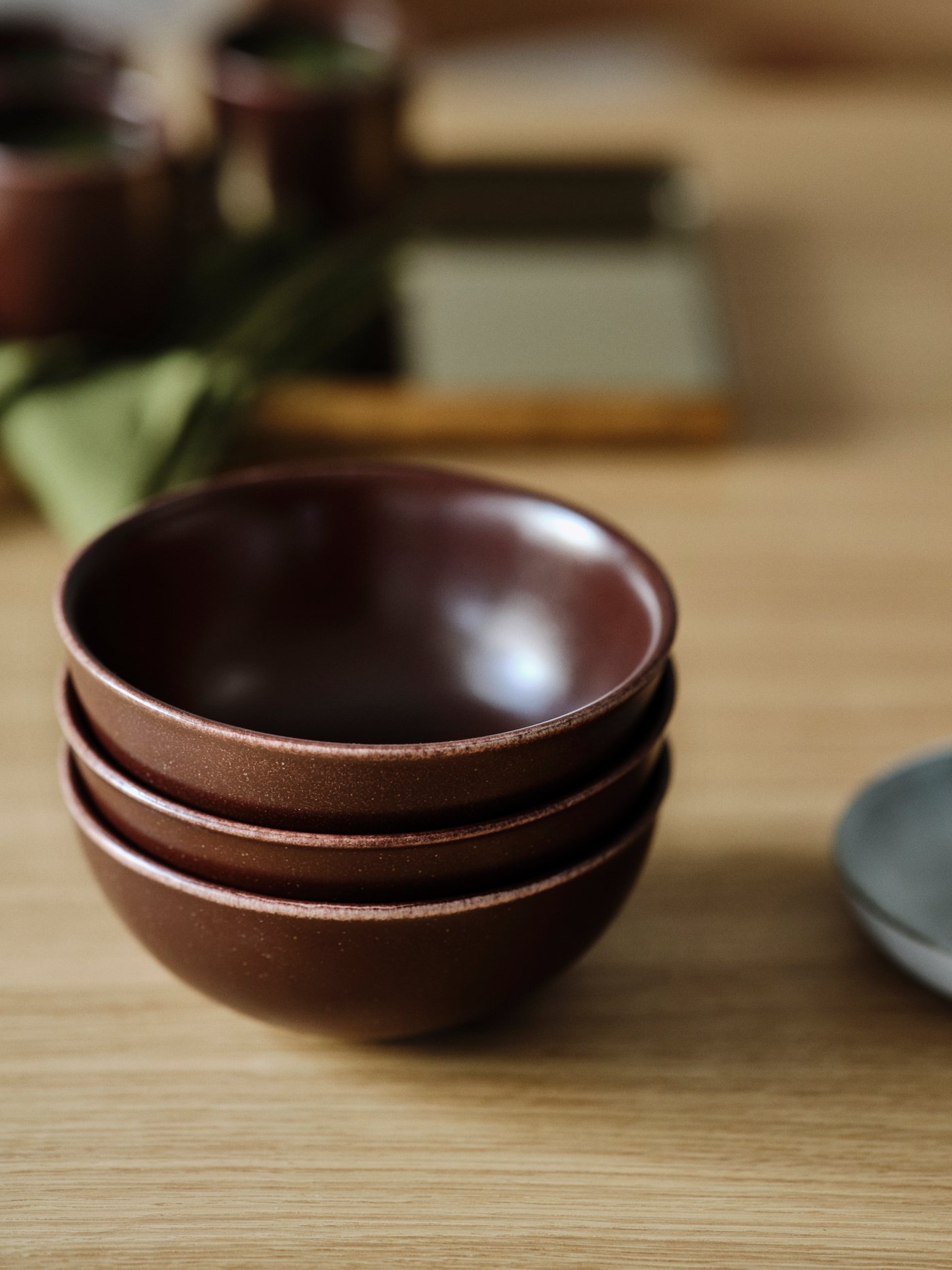 A tabletop with three stacked brown, Japanese-style STOCKHOLM 2025 bowls next to a rustic grey serving plate.