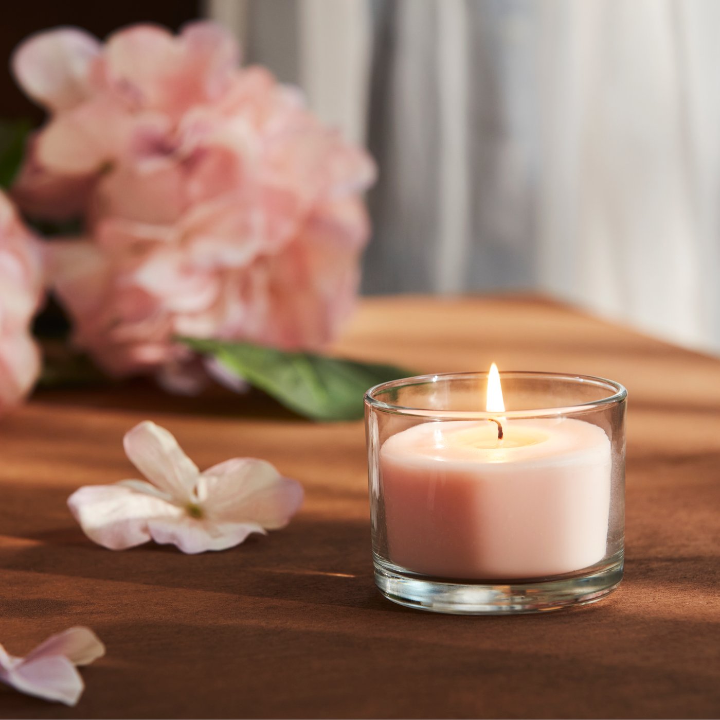 A close-up of a pale pink LOTSFÅGEL scented candle in glass placed on a table with pink flowers in the background.