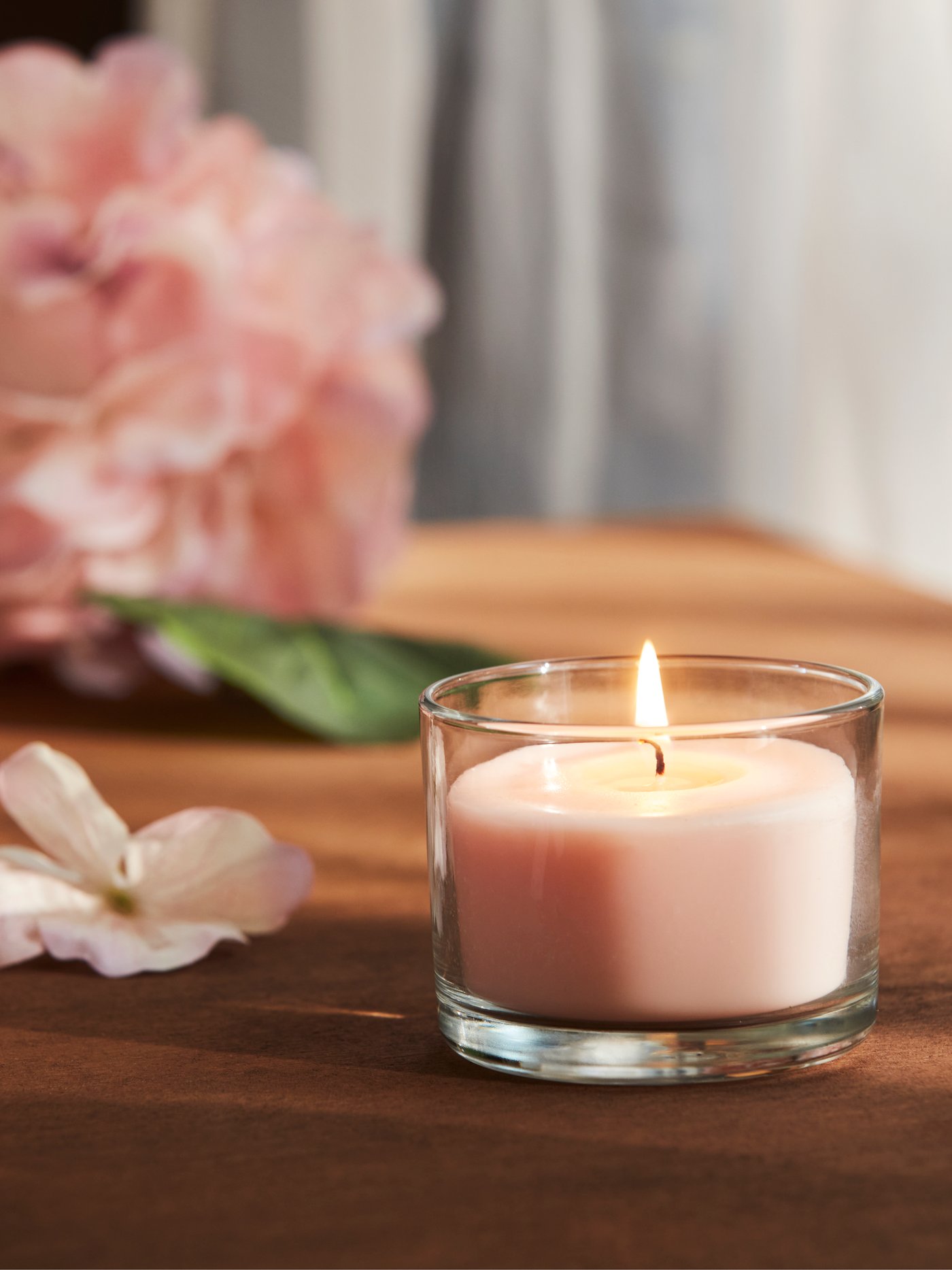 Un primo piano di una candela profumata LOTSFÅGEL rosa pallido in vetro posta su un tavolo con fiori rosa sullo sfondo.