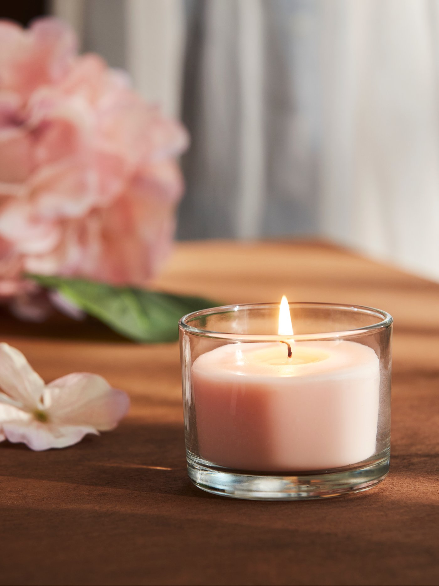 Nahaufnahme einer blassrosa LOTSFÅGEL Duftkerze im Glas, die auf einem Tisch steht, mit rosa Blumen im Hintergrund.