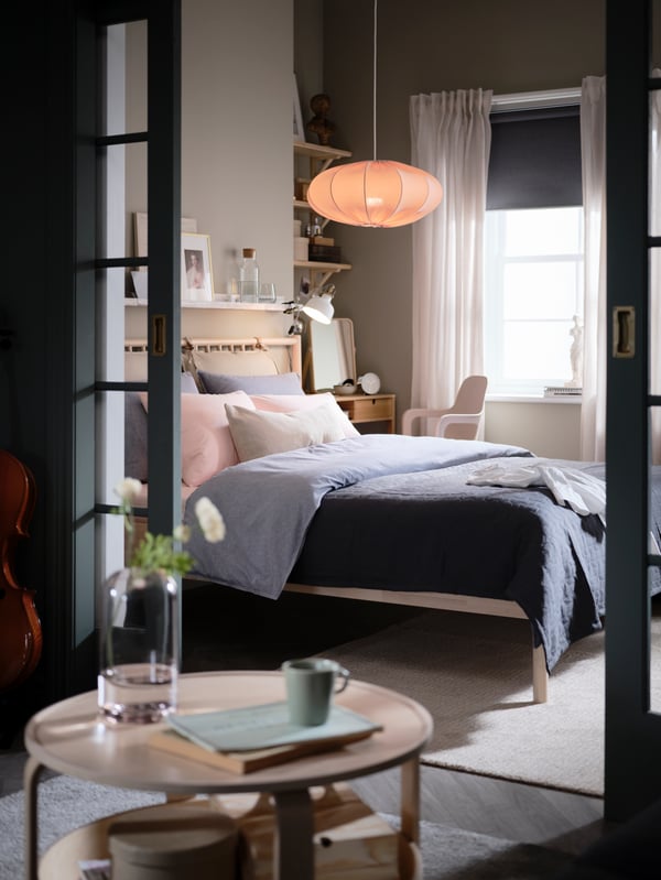 Parted glass doors open onto a bedroom with a BJÖRKSNÄS birch bed, a dressing table and chair, and an oval pendant lamp.