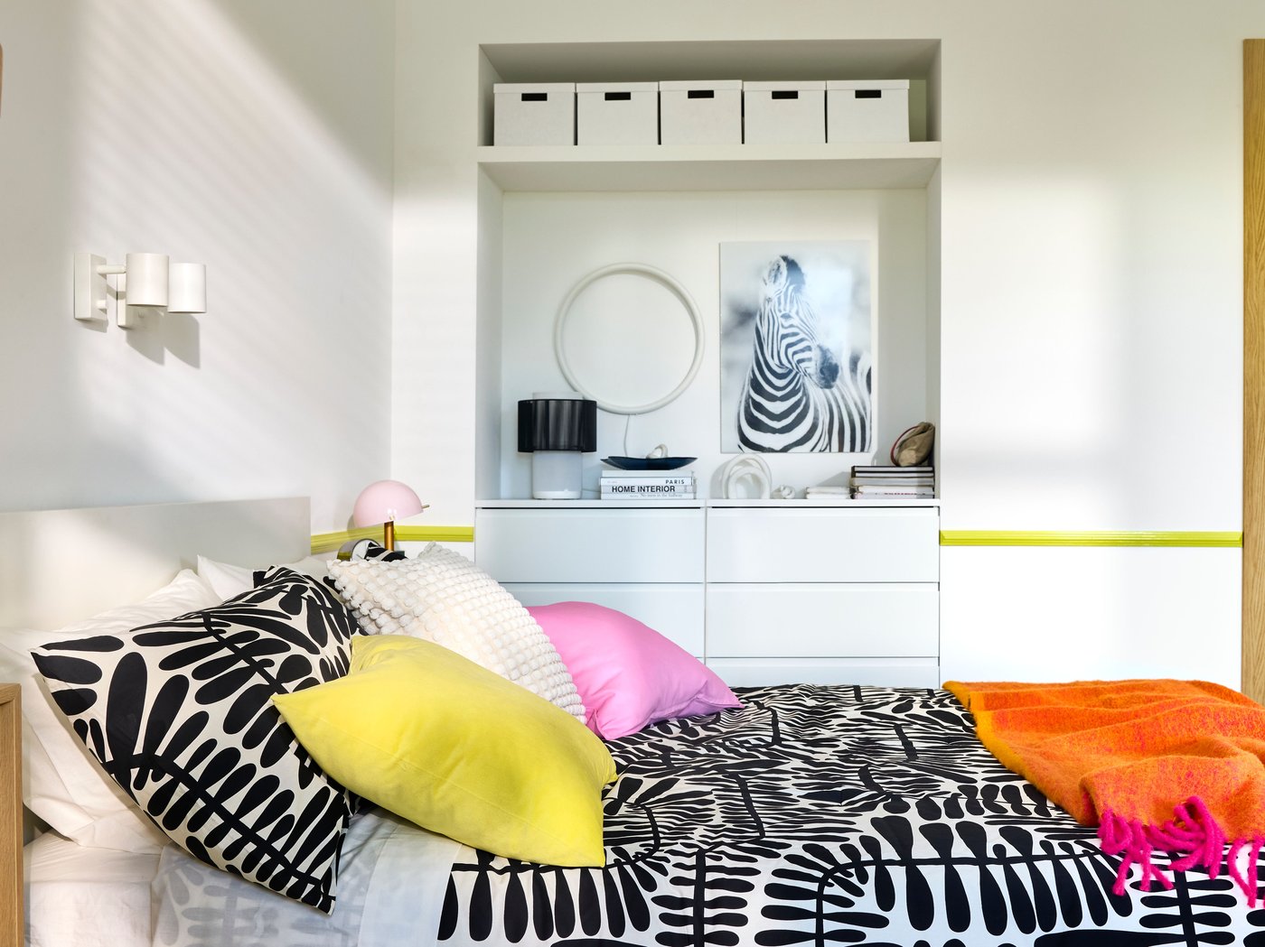 A bedroom with white walls, built-in storage in the back and a double bed with bright pink and black/white bedding.