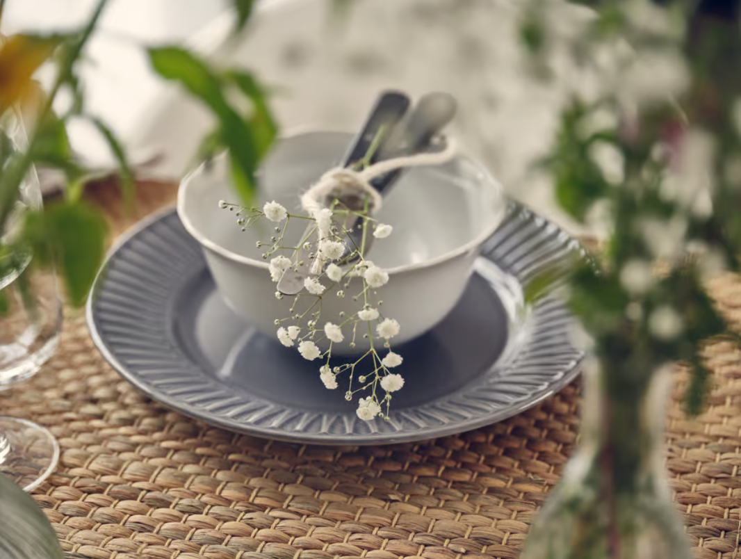 Gestapeltes Geschirr mit Besteck in einer weissen Schale, dekoriert mit zarten weissen Blüten auf geflochtener Platzmatte.