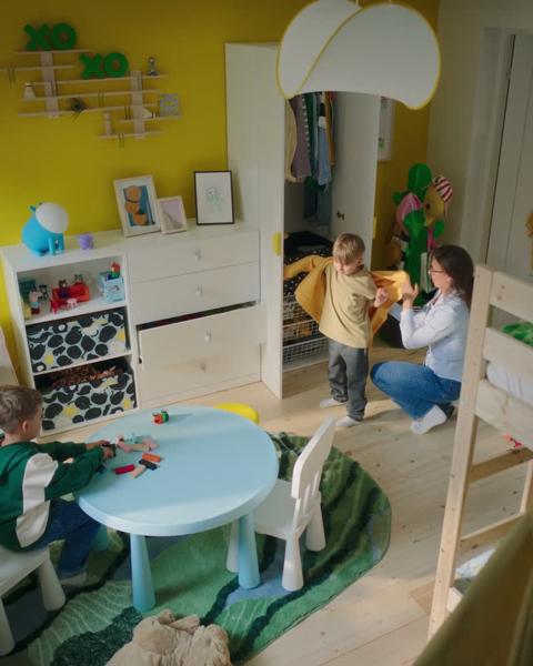 In een levendige gele IKEA kinderkamer helpt een moeder haar zoon in een geel jasje bij een witte kledingkast, terwijl een ander kind speelt aan een IKEA LÄTT tafel en stoelen onder een opvallende IKEA KRUSNING lampenkap.