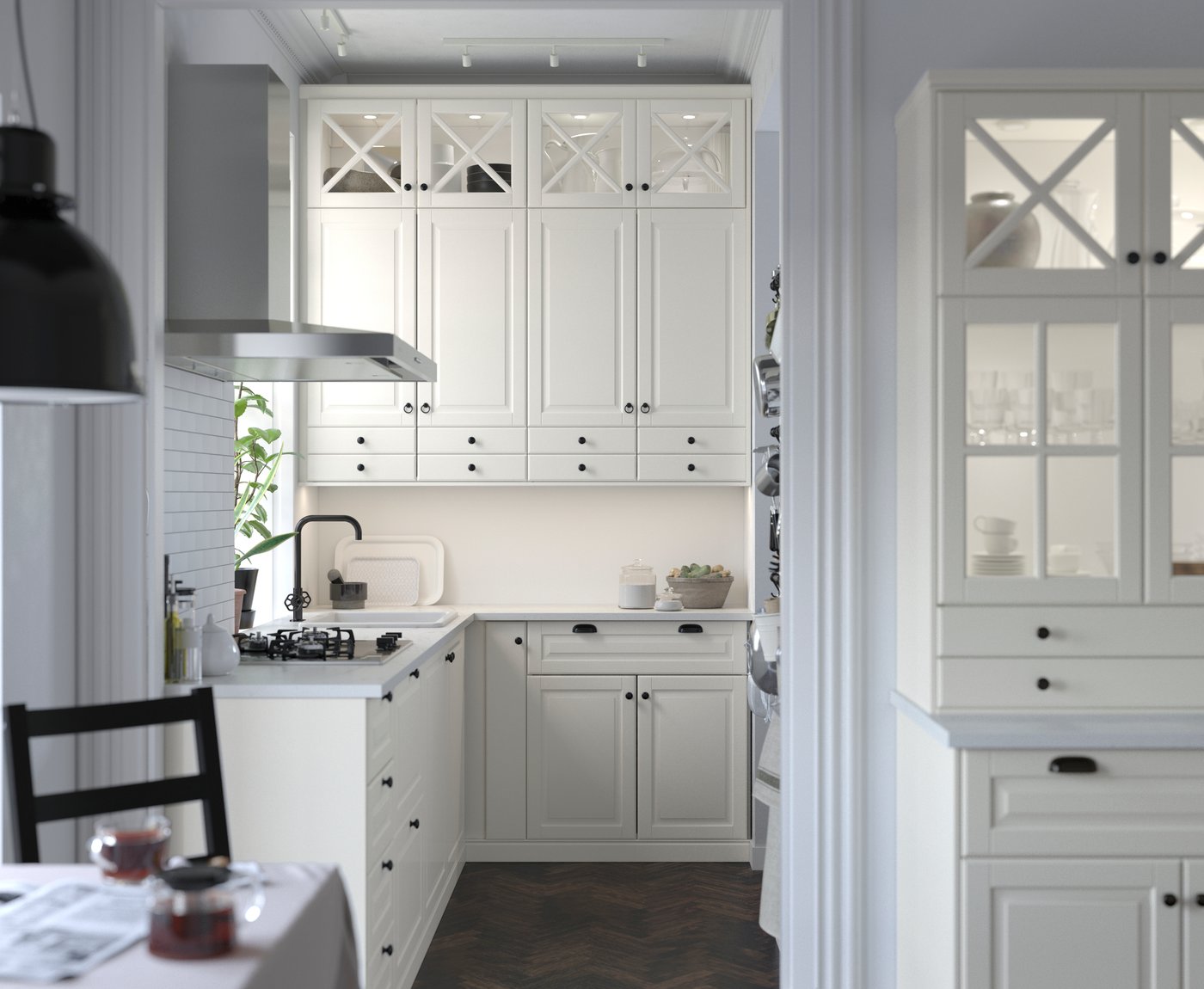 Bucătărie modernă și luminoasă cu dulapuri albe, mânere negre, chiuvetă, aragaz cu hotă de inox, dulap vitrină cu veselă și podea din lemn închis la culoare.