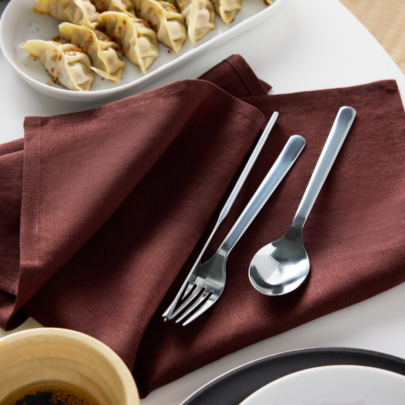 A fork, a knife and a spoon from the IKEA 365+ cutlery series lie on a dark red STOCKHOLM 2025 napkin.